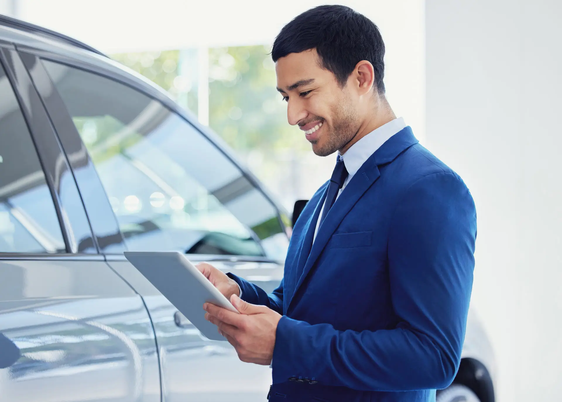 Car sales person smiling down at a tablet while standing next to a car
