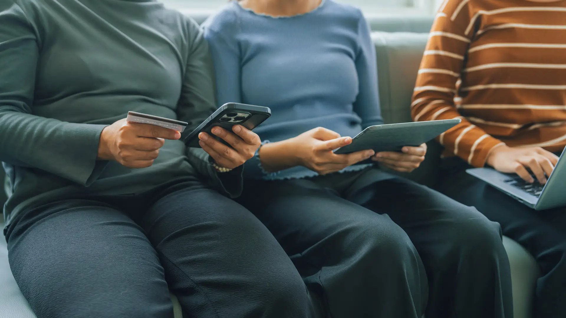 Three people on a couch. One is holding a debit card and their phone, one is holding a tablet, and one is holding a laptop.